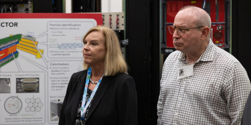 A woman and man stand and look at a person speaking.